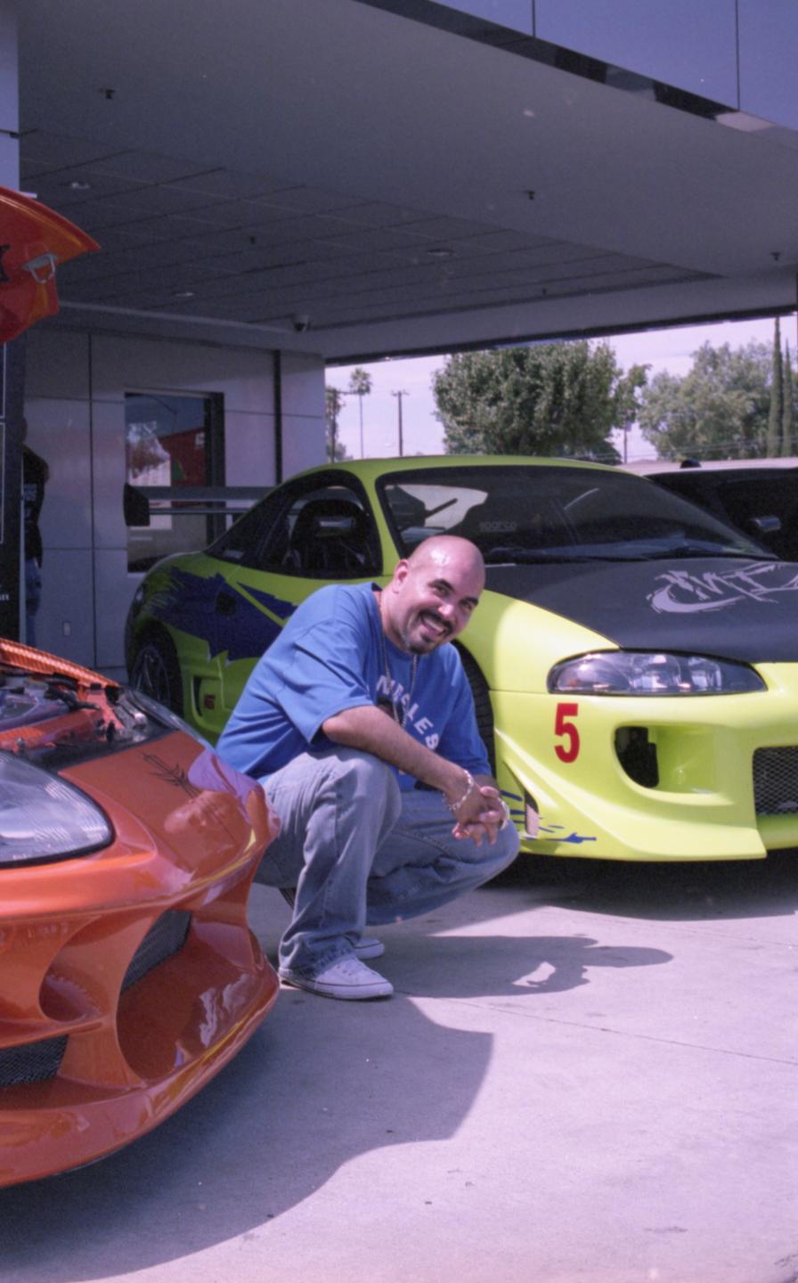 A man crouches next to a bright yellow sports car with an orange sports car in the foreground.