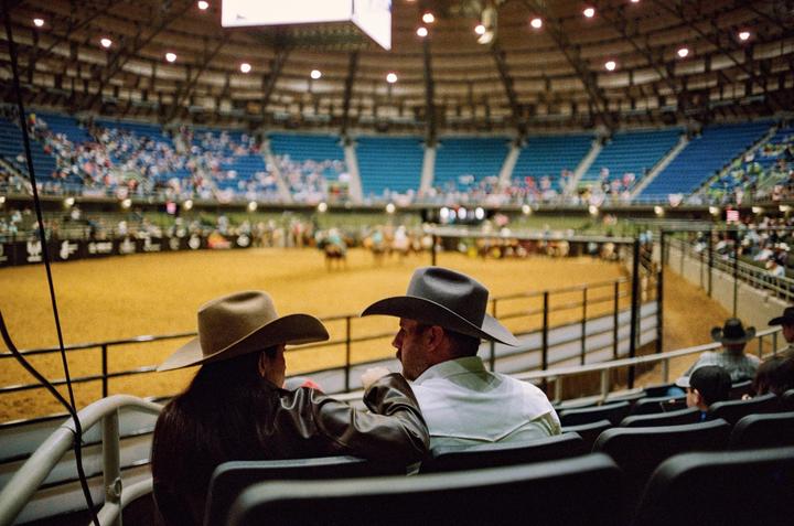 A couple wearing cowboy hats watches a rodeo from the stands of an arena.