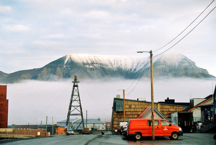 A red van is parked on a street with buildings and a large snowy mountain partially obscured by fog in the background.