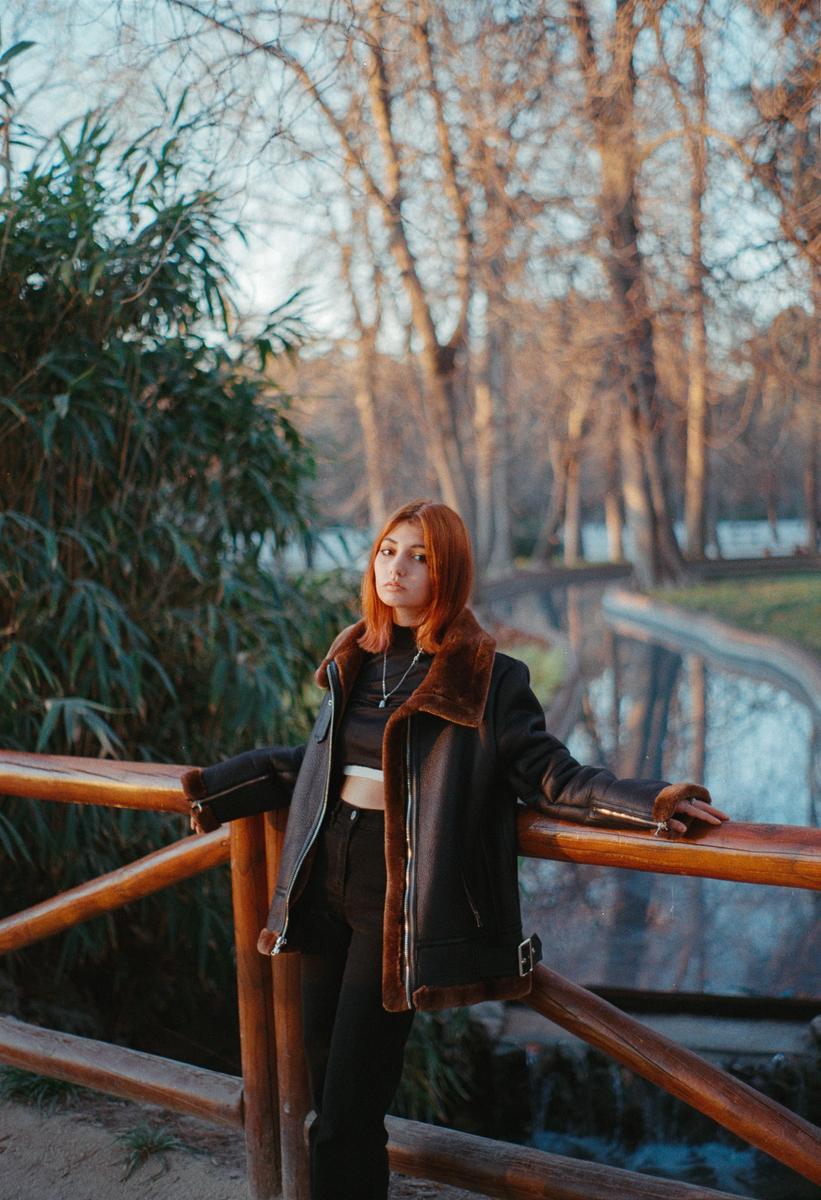 A woman with red hair and a black jacket stands by a wooden fence next to a body of water with trees in the background.