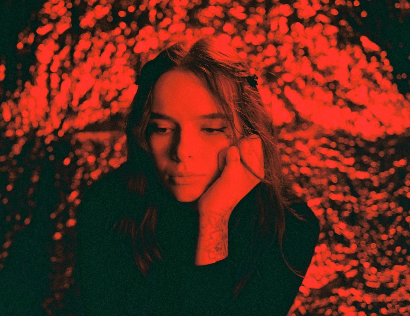 A woman with her hand on her chin looks down against a red, blurry, bokeh background.