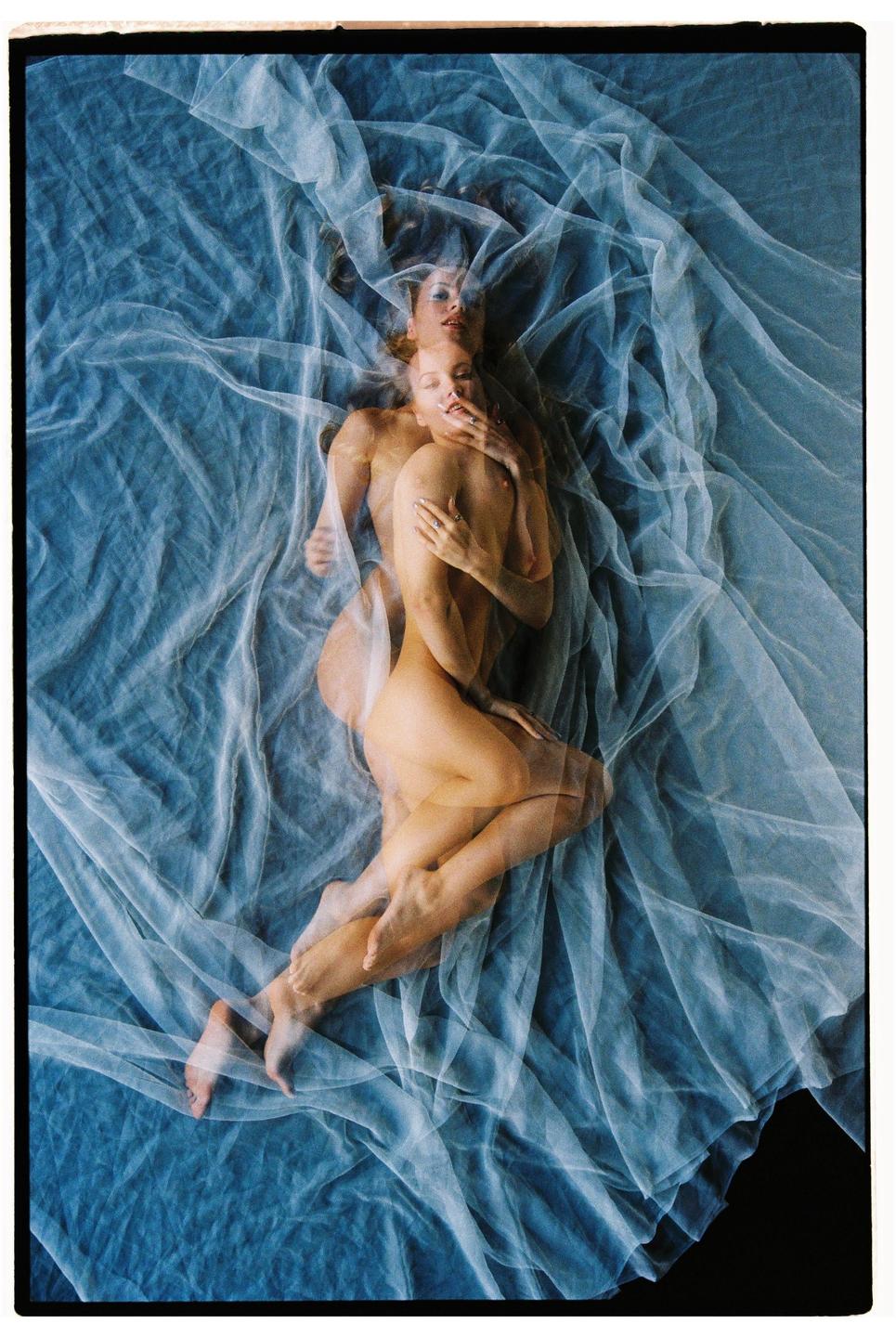 Two women are intertwined and covered by a sheer white fabric on a blue surface.