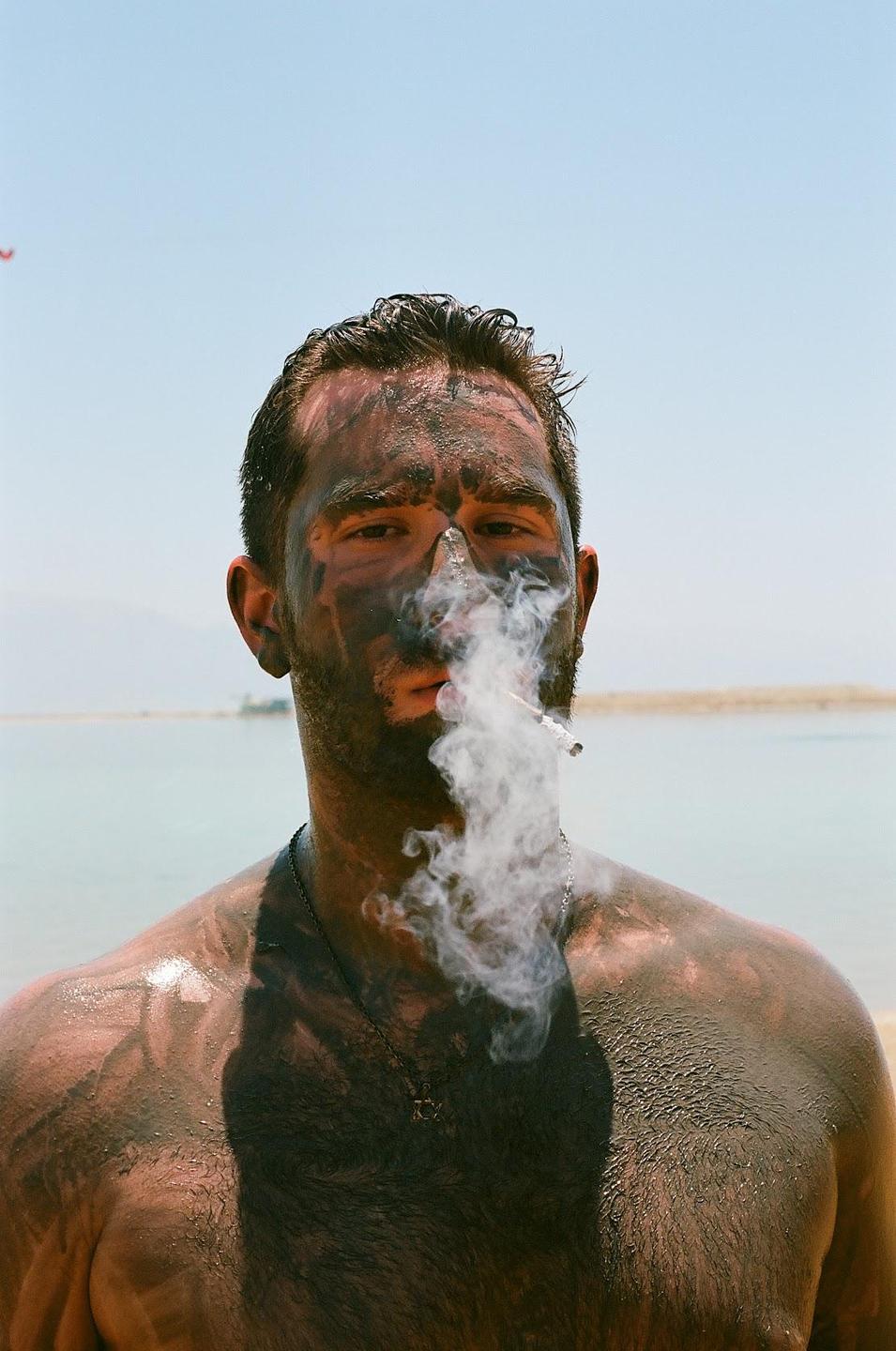 A man with mud on his face and body exhales smoke from a cigarette while standing in water.