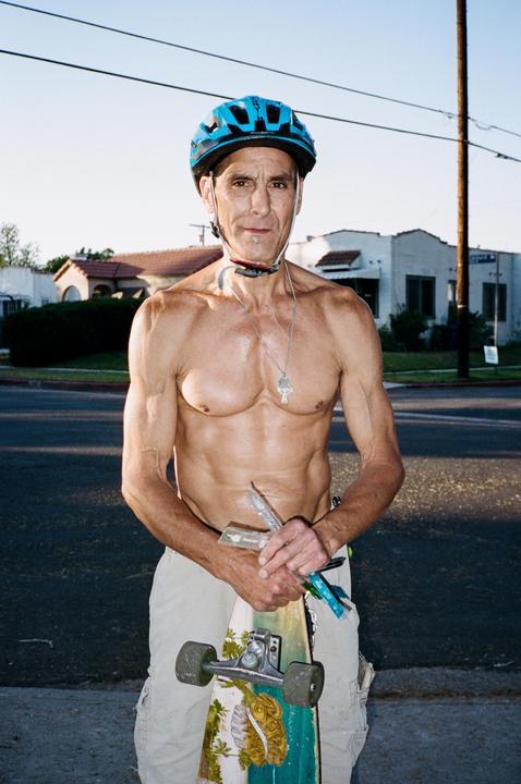 A shirtless man wearing a blue helmet and holding a skateboard stands on a street.