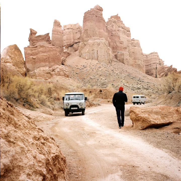 A man in a red hat walks away from a white van on a dirt road toward rock formations.