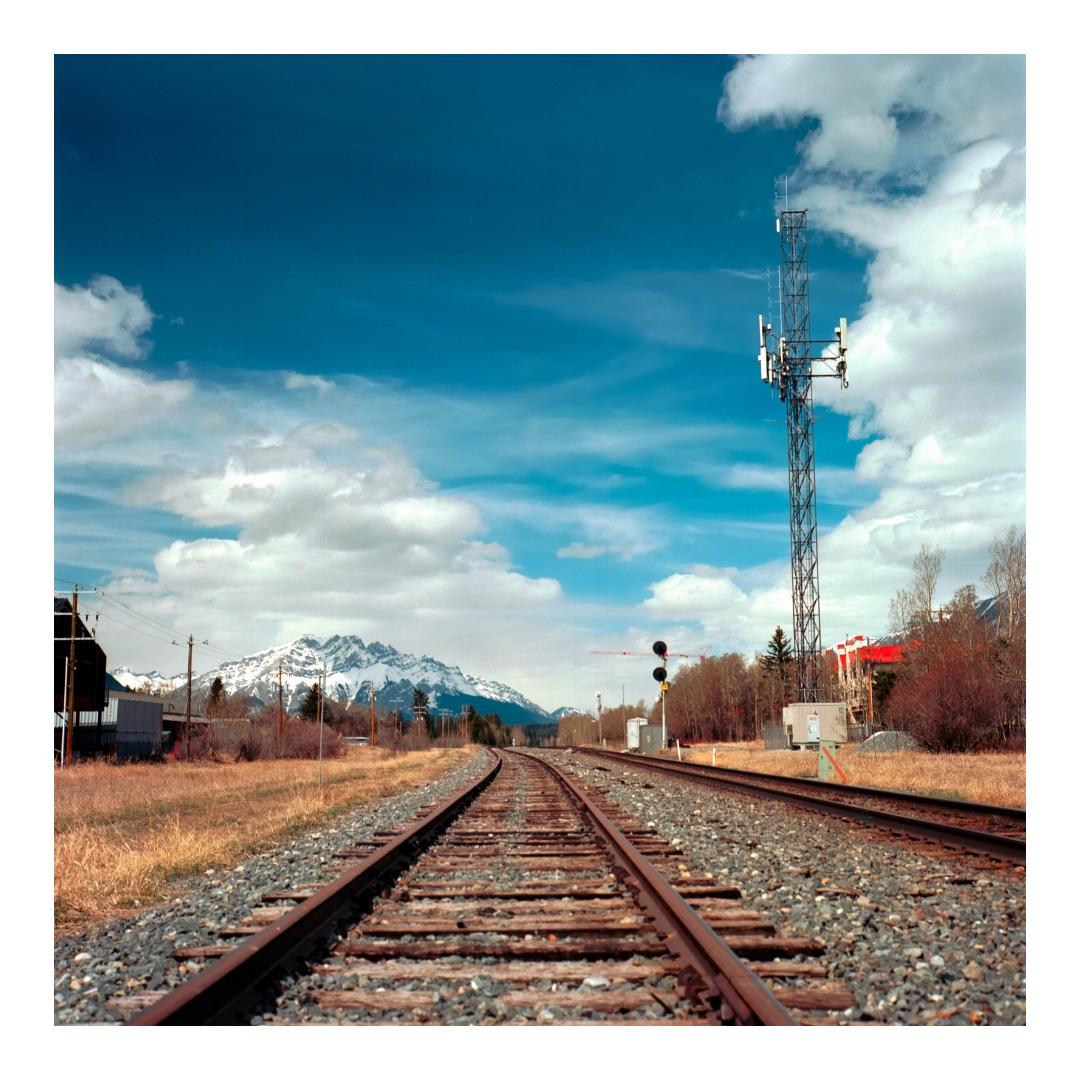 Two parallel railroad tracks curve into the distance toward a snow-capped mountain range under a blue sky with white clouds.