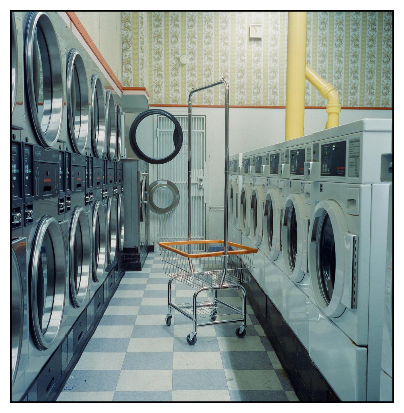 A laundry cart stands in the middle of a laundromat with rows of washing machines and dryers.