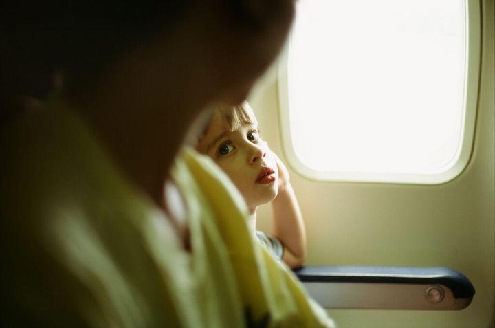 A child with light hair looks over the shoulder of another person, with a bright window visible to the right.