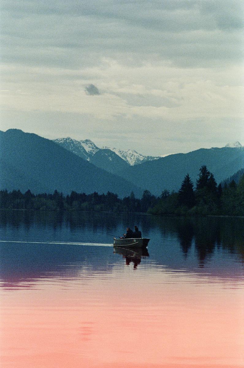 Two people in a small boat move across a lake with mountains and trees in the background.