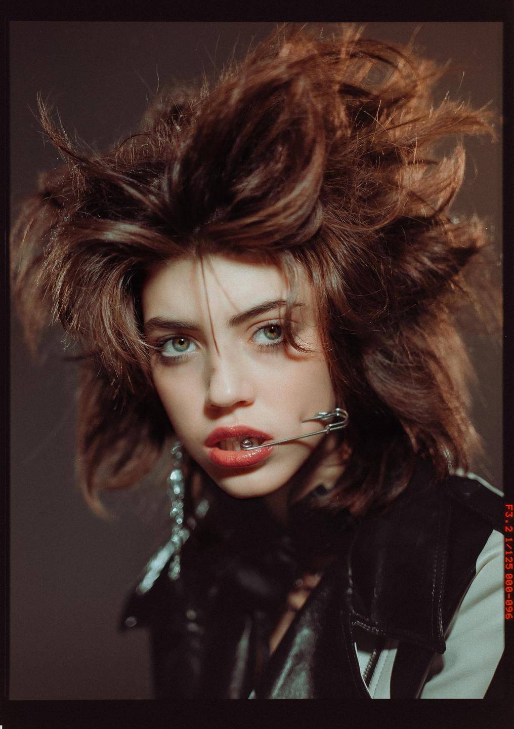 A woman with messy hair and a safety pin in her mouth looks directly at the viewer.