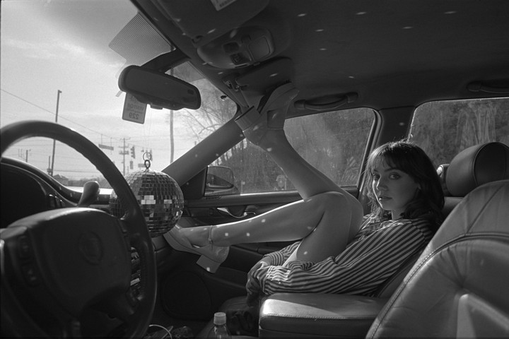 A woman with her legs up is sitting in a car with a disco ball hanging from the rearview mirror.