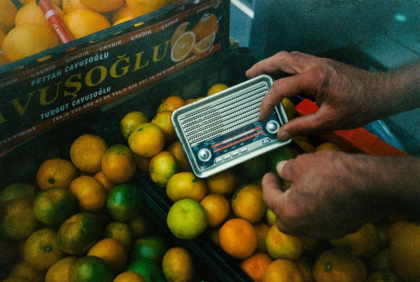 A person's hands hold a small radio over a crate of oranges and tangerines.
