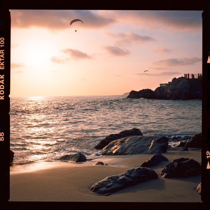 Two paragliders fly over a rocky beach at sunset.