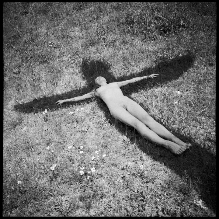 A nude woman lies on her back in a grassy field with her arms outstretched, casting a shadow.