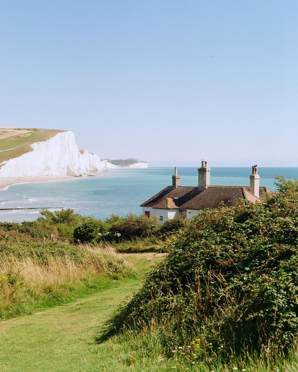 A white house with a brown roof is nestled among green bushes and grass, overlooking a beach with white cliffs and blue water.