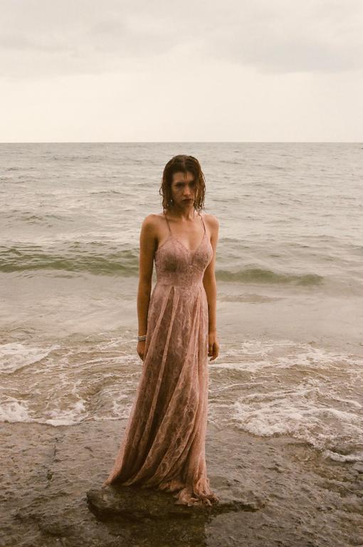 A woman in a long pink dress stands in the shallow water of a beach with waves breaking around her feet.