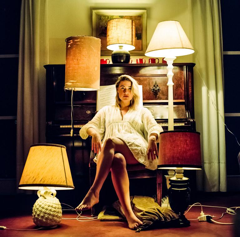A woman sits in a chair in front of a piano, surrounded by lamps.