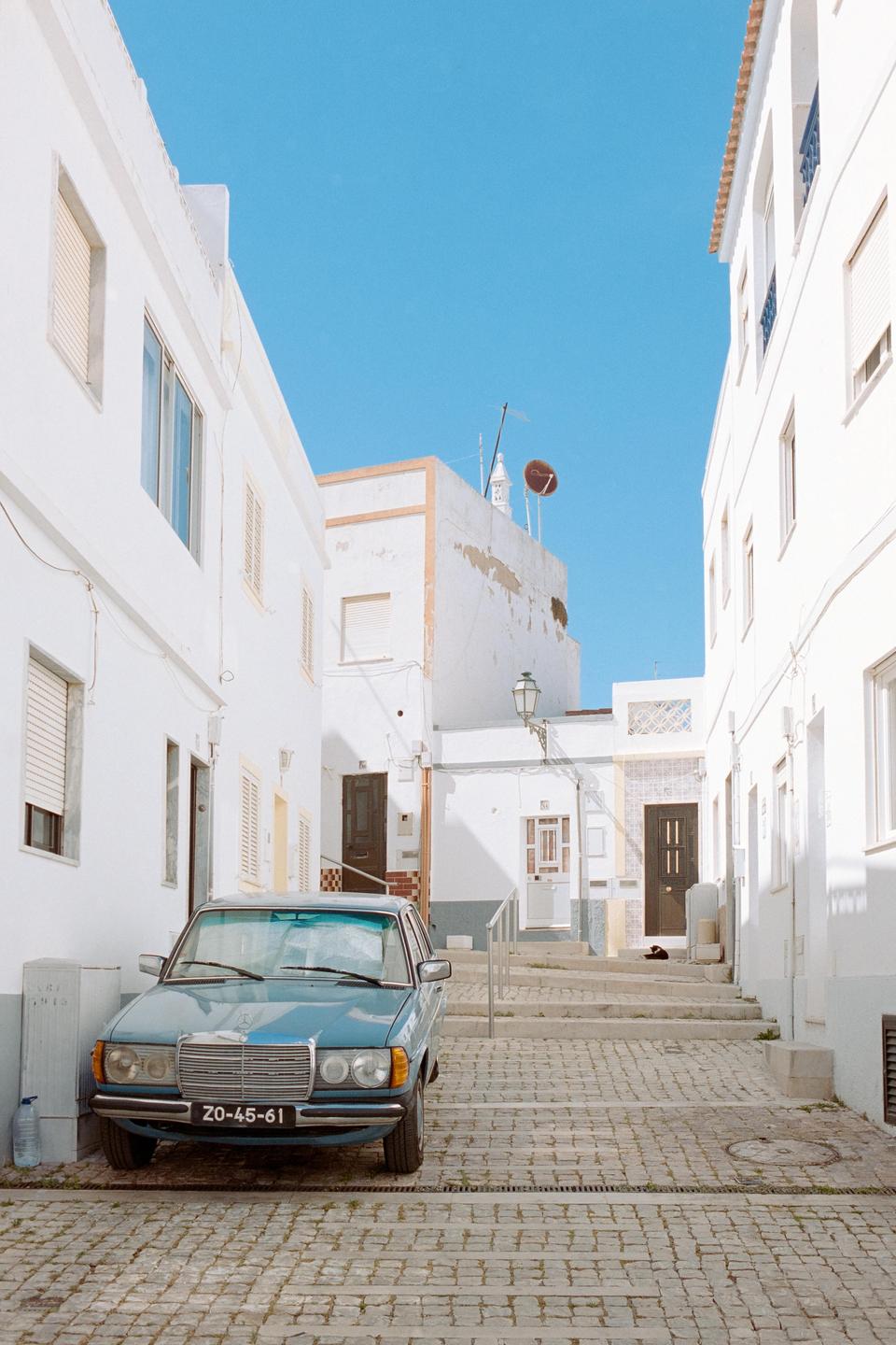 A blue car is parked on a cobblestone street between white buildings under a clear blue sky.