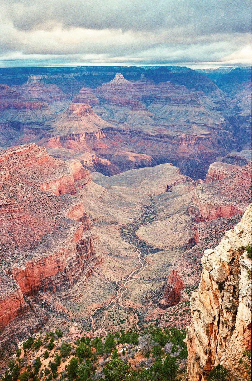 A wide view of the Grand Canyon shows a winding path through the canyon floor and dramatic rock formations under a cloudy sky.