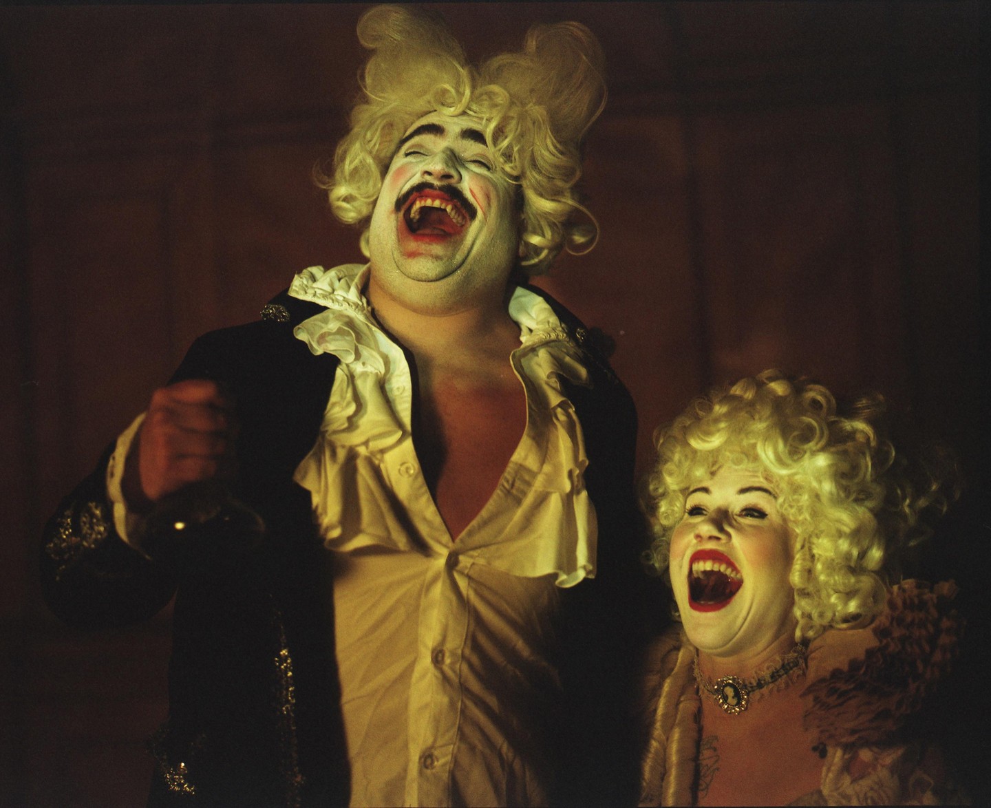 A man and a woman in theatrical costumes and wigs laugh with their mouths wide open.