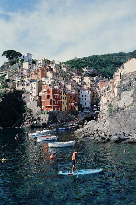 A man paddleboards in a harbor with colorful buildings on a hillside in the background.