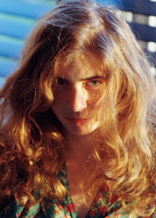 A woman with long, wavy hair looks directly at the viewer, with sunlight illuminating her hair and face.