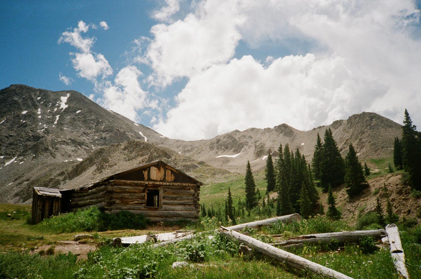 A wooden cabin sits in a grassy field with mountains and pine trees in the background under a blue sky with clouds.