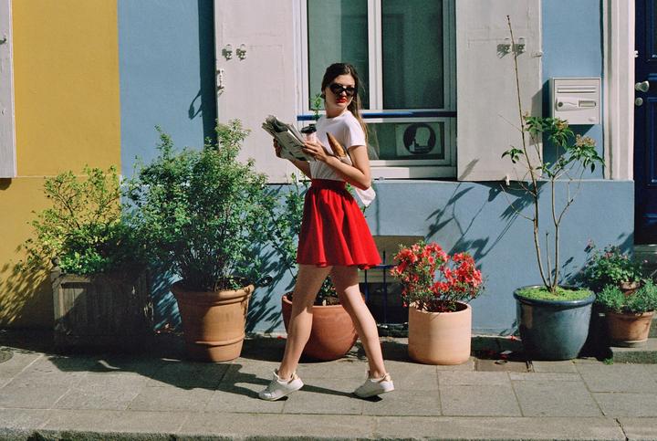 A woman in a red skirt walks past colorful buildings and potted plants while holding a newspaper, coffee, and baguette.