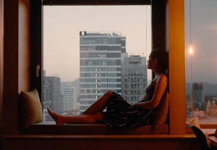 A woman sits on a window seat looking out at a city skyline at dusk.