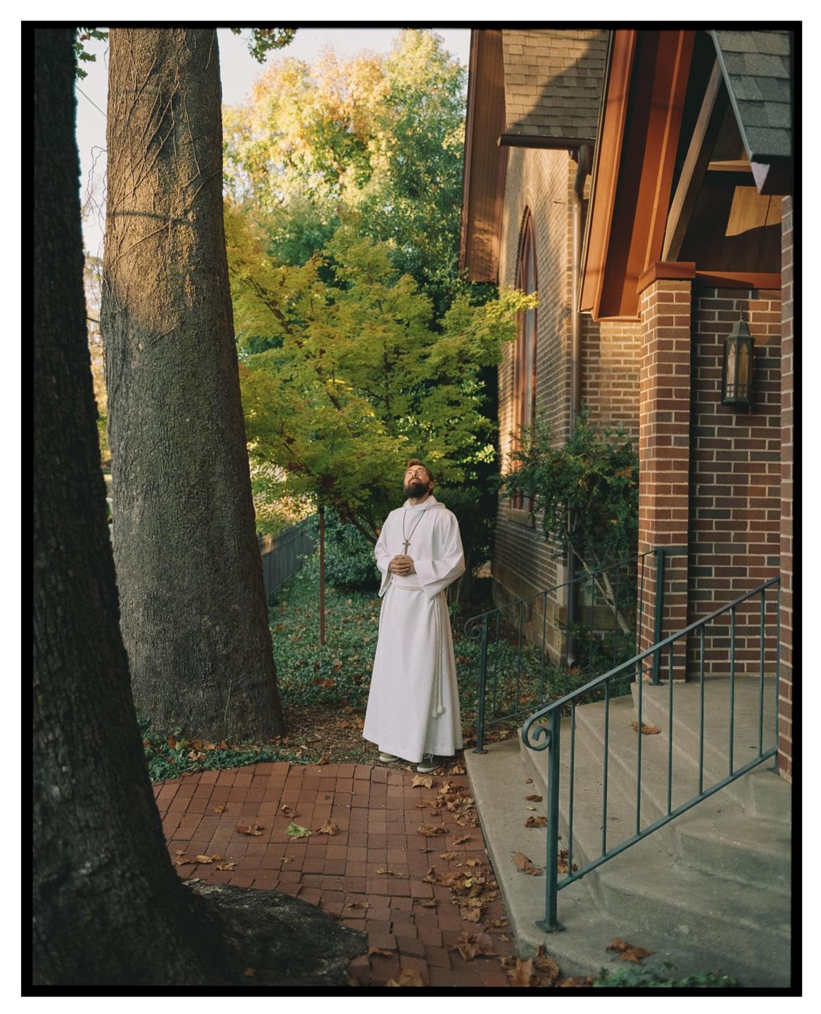 A man in a white robe stands on a brick path next to a building with trees in the background.