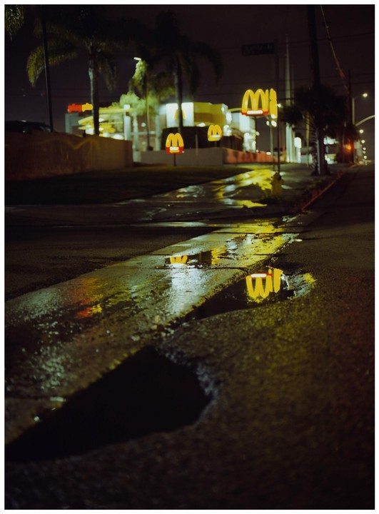 Golden arches from a McDonald's are reflected in puddles on a wet street at night.