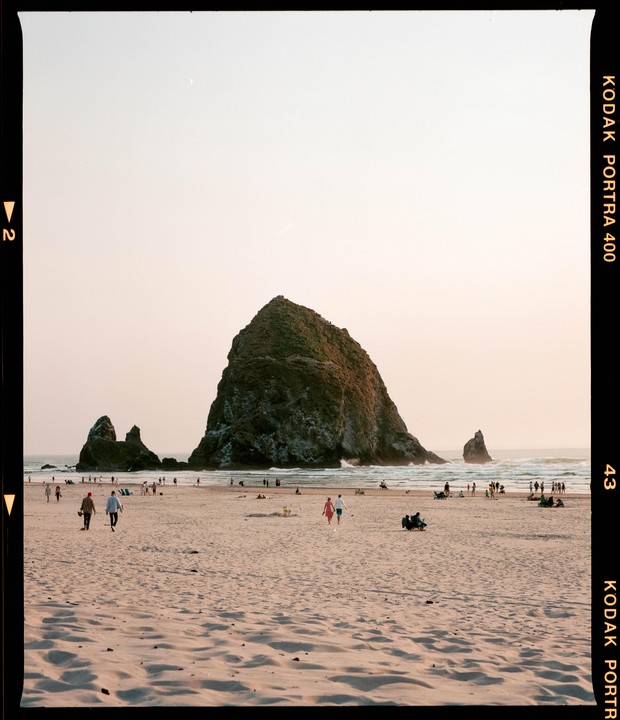 A large rock formation stands on a sandy beach with people walking and sitting near the ocean.