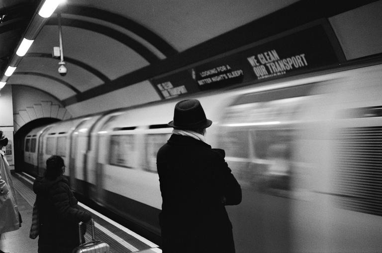 A man in a hat stands on a subway platform as a train speeds by.
