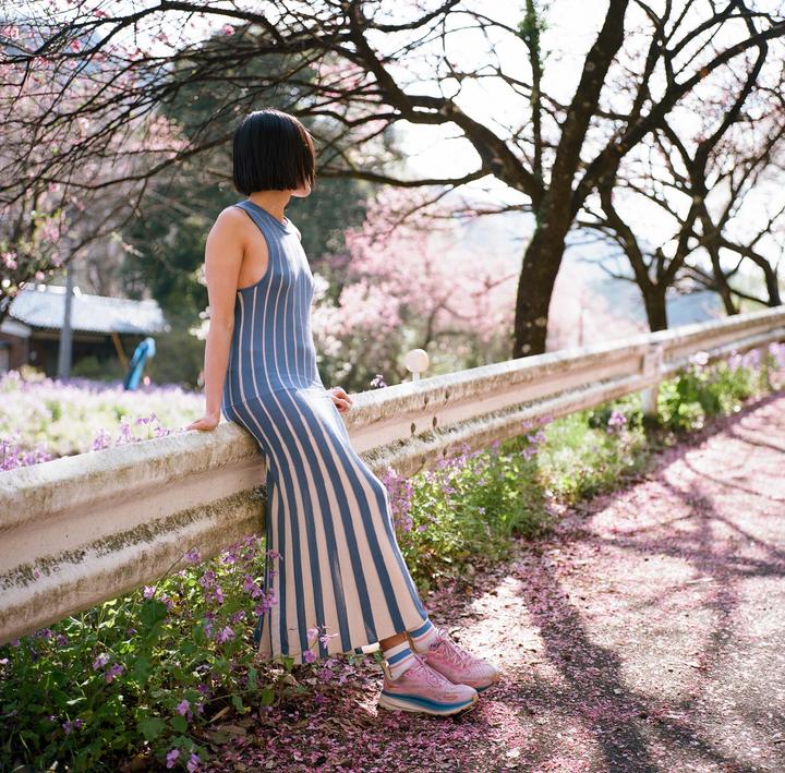 A woman in a striped dress sits on a guardrail next to a road covered in pink petals.