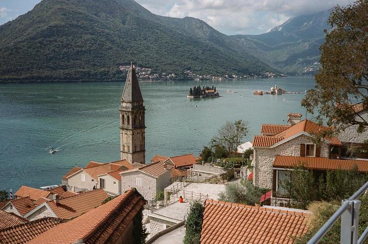 A tall bell tower rises above terracotta rooftops next to a bay with mountains in the background.