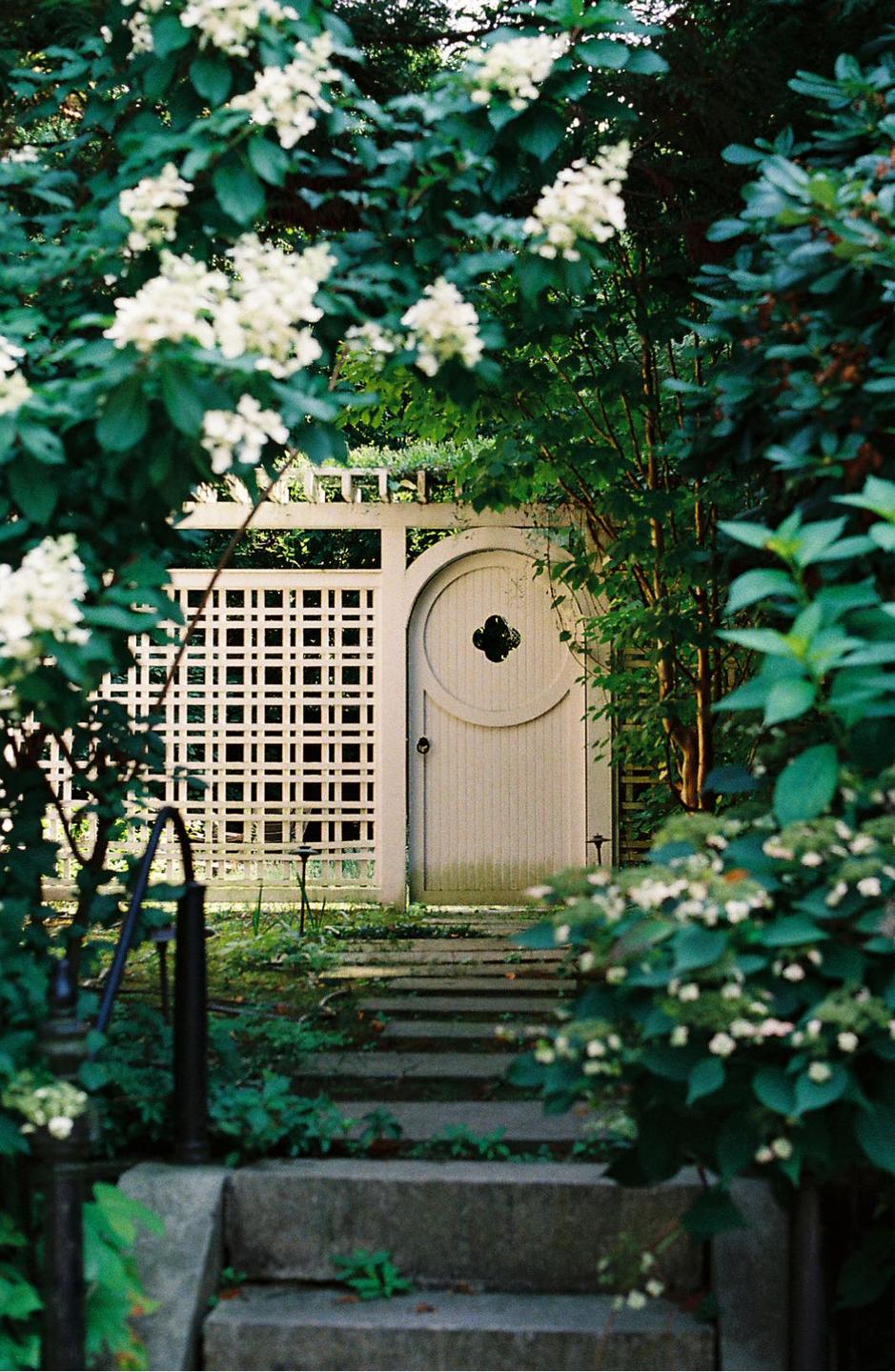 A white garden gate with a circular design is framed by green foliage and white flowers, with stone steps leading up to it.