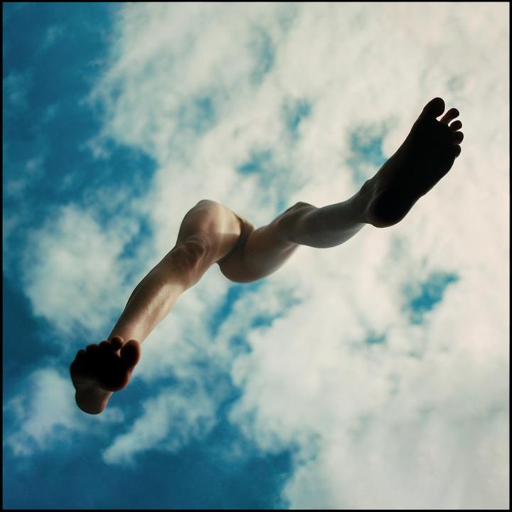 A person's legs and feet are visible against a blue sky with white clouds, as if falling or flying.