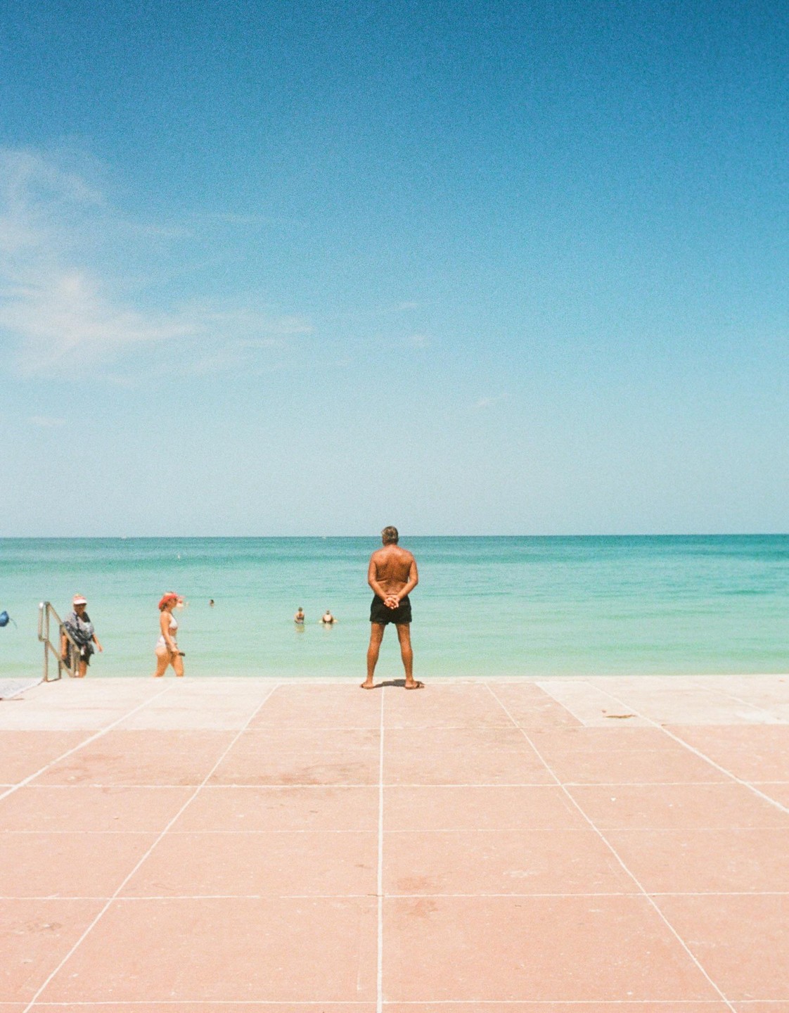 A man stands on a tiled surface looking out at the ocean where other people are swimming.