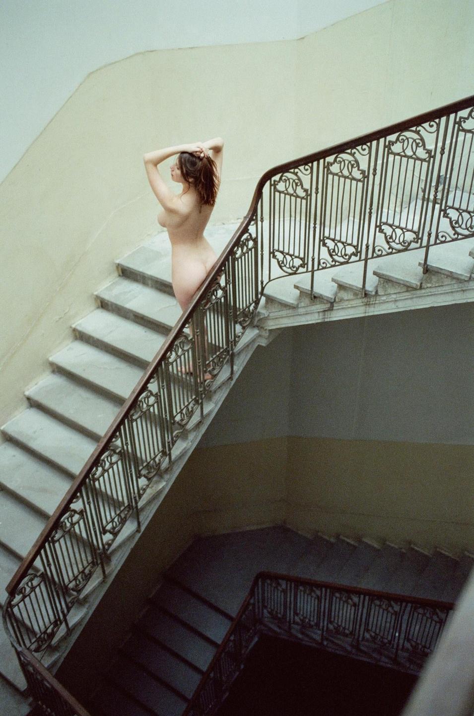 A nude woman stands on a marble staircase with an ornate railing, looking away from the camera.