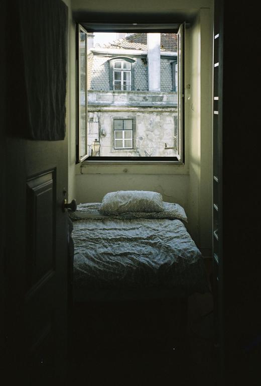 A bed with a patterned duvet and pillow is positioned beneath an open window looking out onto old buildings.