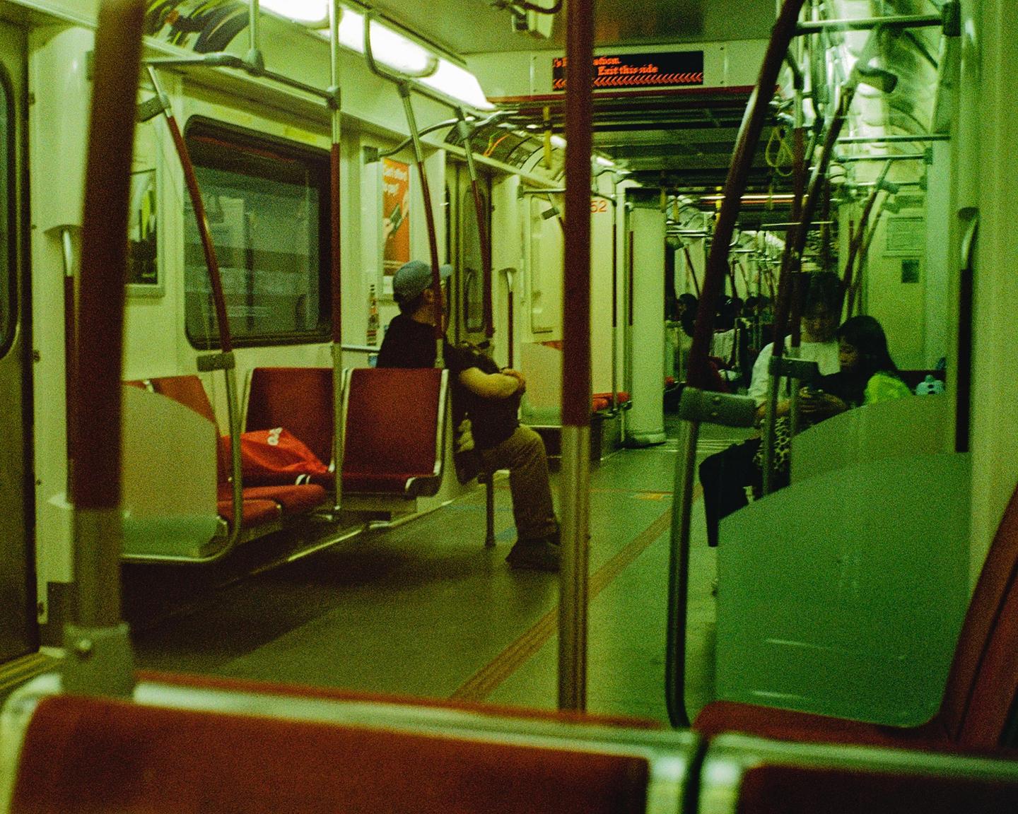 People are riding a subway car with red seats and green-tinted lighting.