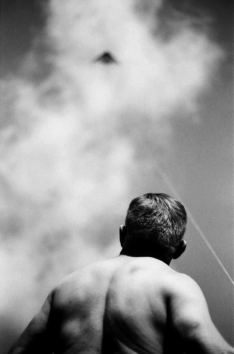 A man with his back to the viewer looks up at a kite flying in a cloudy sky.