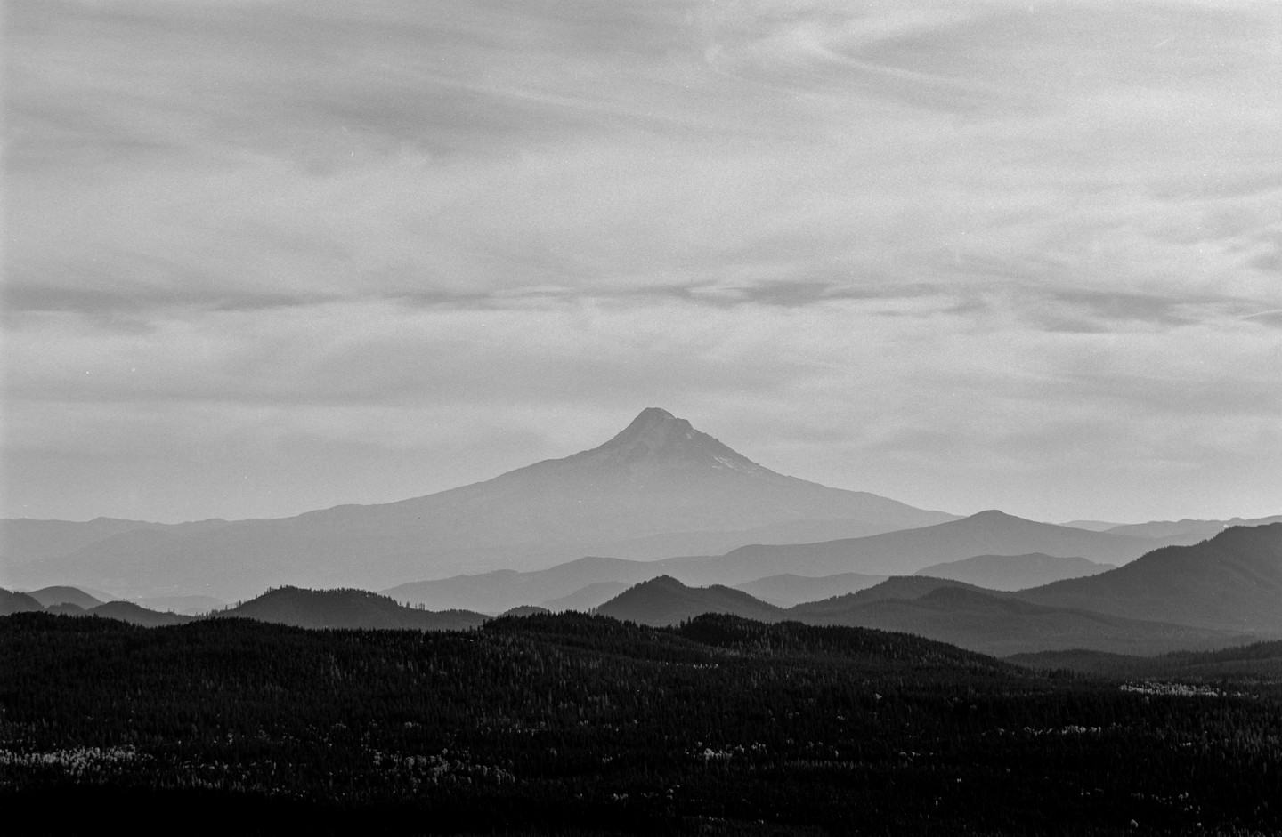 A large mountain peak rises above several layers of forested hills under a cloudy sky.