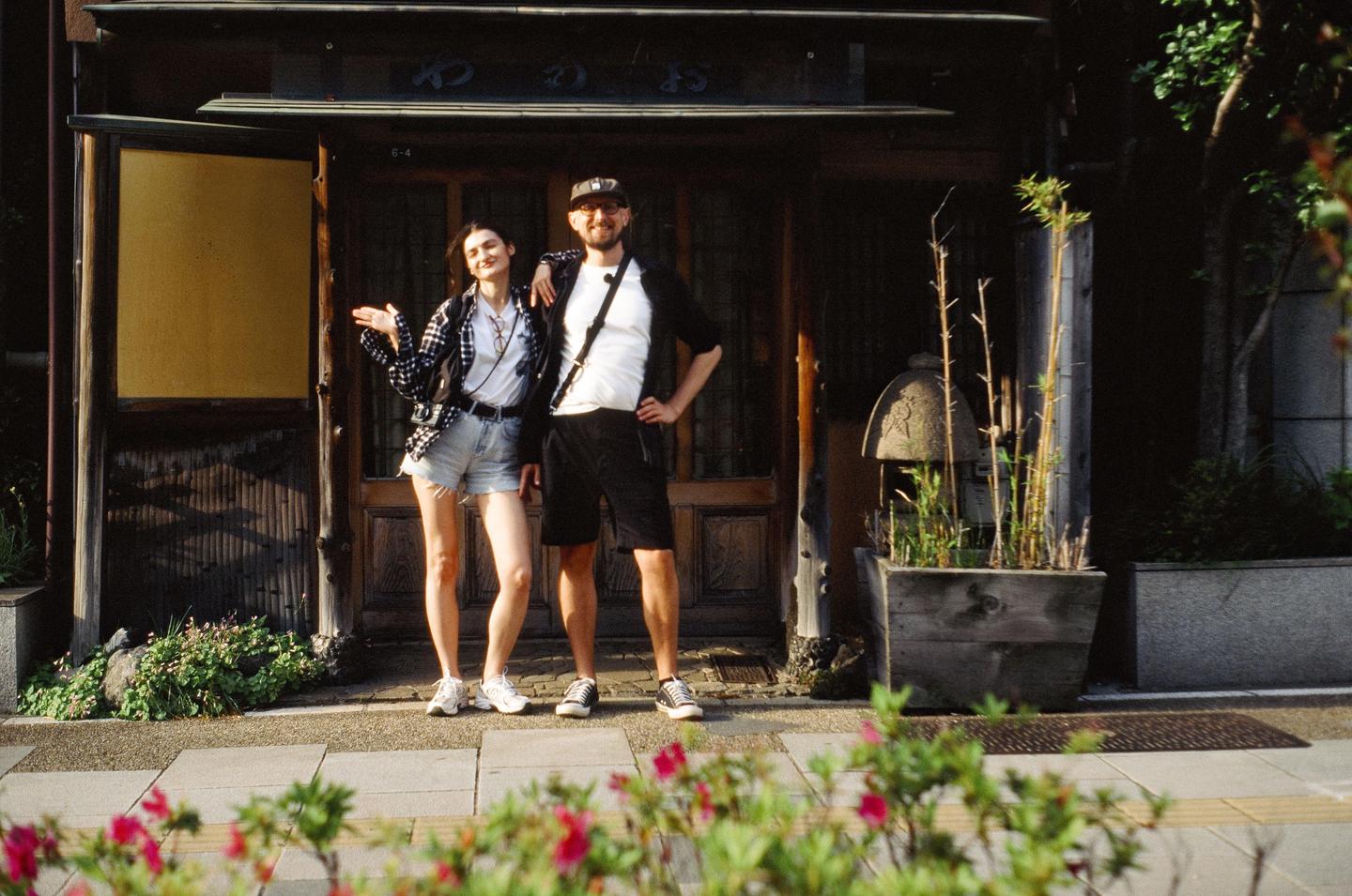 A couple stands in front of a traditional Japanese building with a stone lantern and bamboo plants to their right.