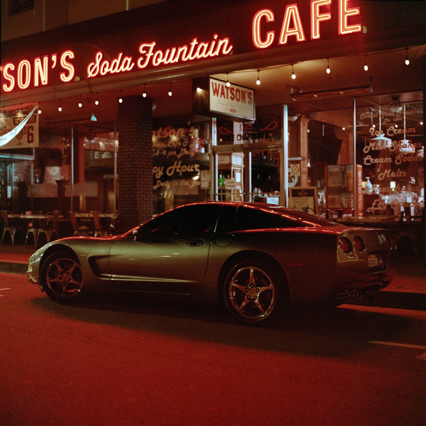A sports car is parked on a street in front of a cafe with neon signs at night.