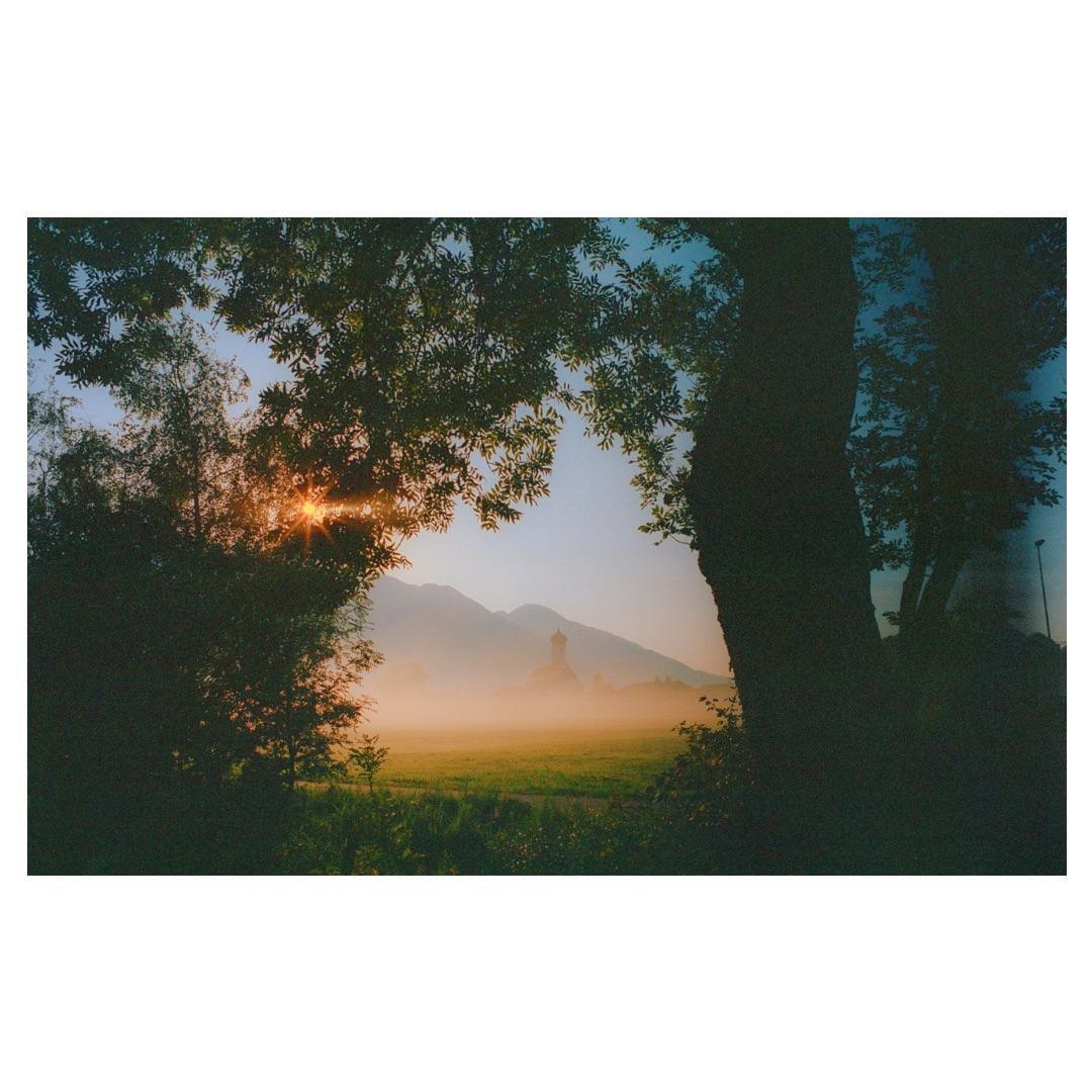 The sun shines through trees onto a hazy field with a church and mountains in the distance.