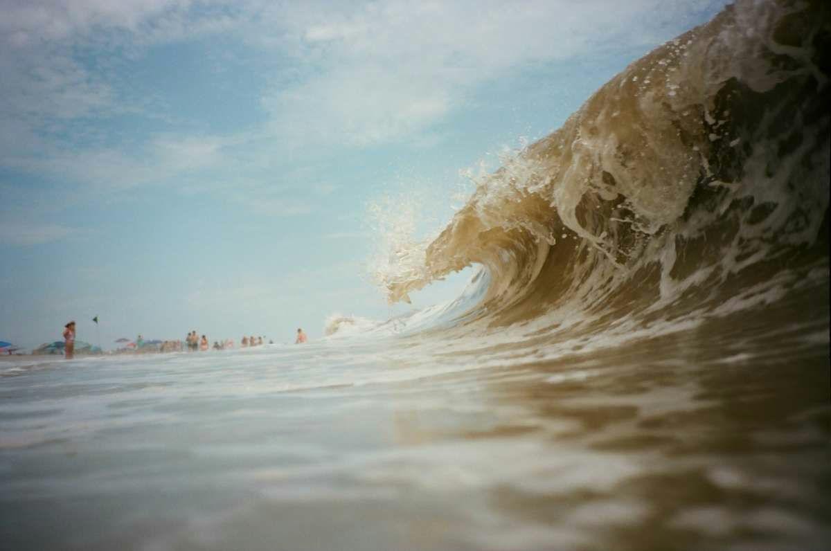 A large wave crashes towards the viewer with people on a beach in the background under a blue sky.