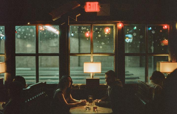 A couple sits at a table in a dimly lit bar with an exit sign and string lights visible through the window.