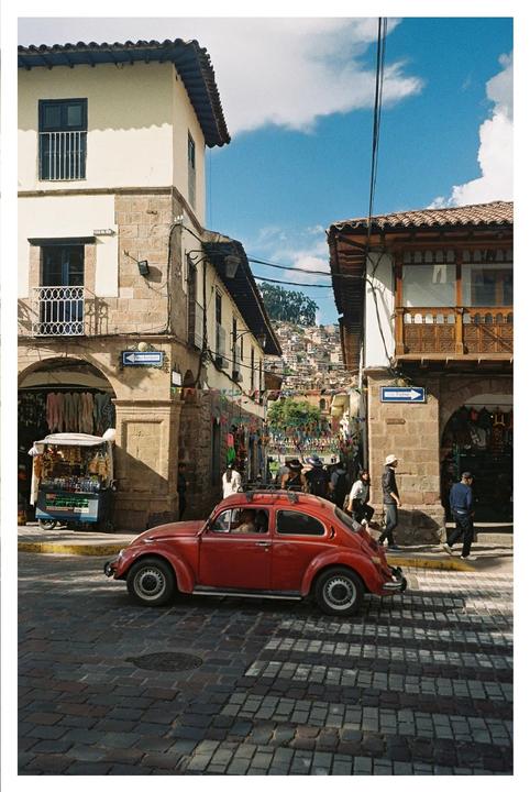 A red car is parked on a cobblestone street in a town with buildings and people.