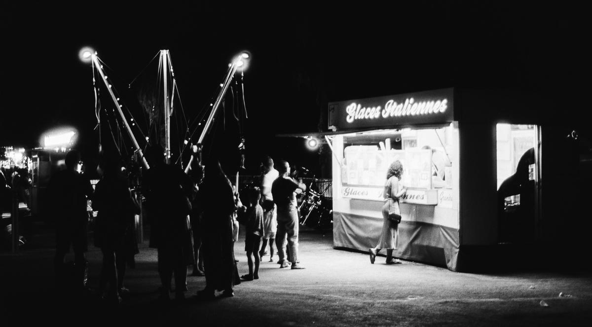 A woman stands at a "Glaces Italiennes" stand at night, with other people and bright lights in the background.